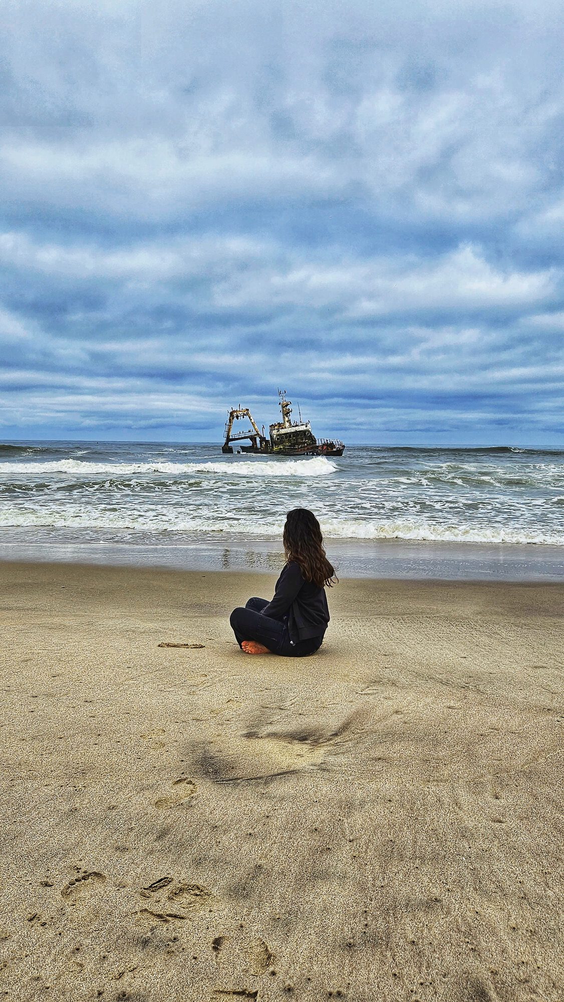 Kelly sitting on the Skeleton Coast beach, a shipwreck visible in the Atlantic behind her