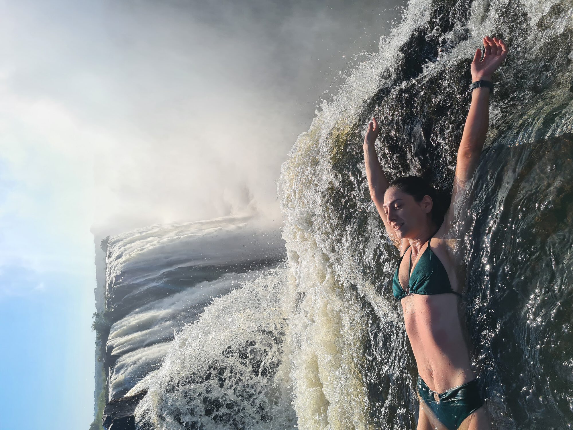 Kelly with arms raised in the Victoria Falls infinity pool, mist rising from the gorge